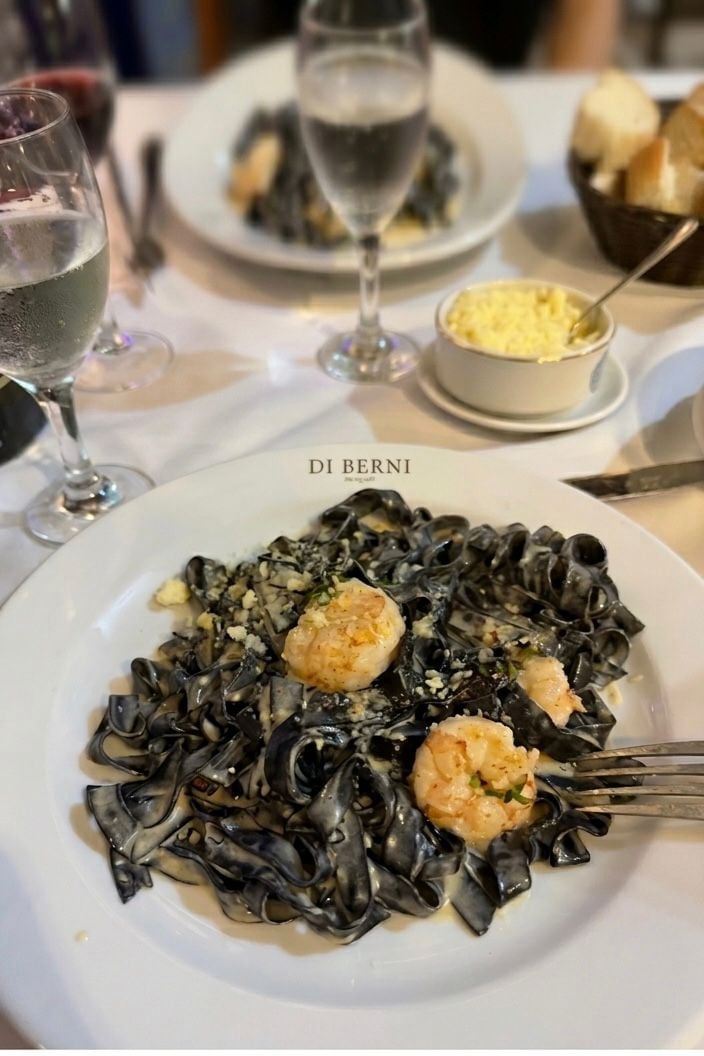 Plate of black squid ink pasta with shrimp at Di Berni restaurant, accompanied by wine glasses and side dishes