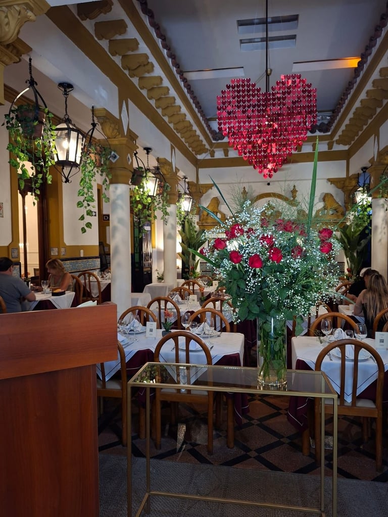 Elegant restaurant interior with wooden chairs, white tablecloths, hanging pink heart decoration, fresh flowers, and decorative plants.