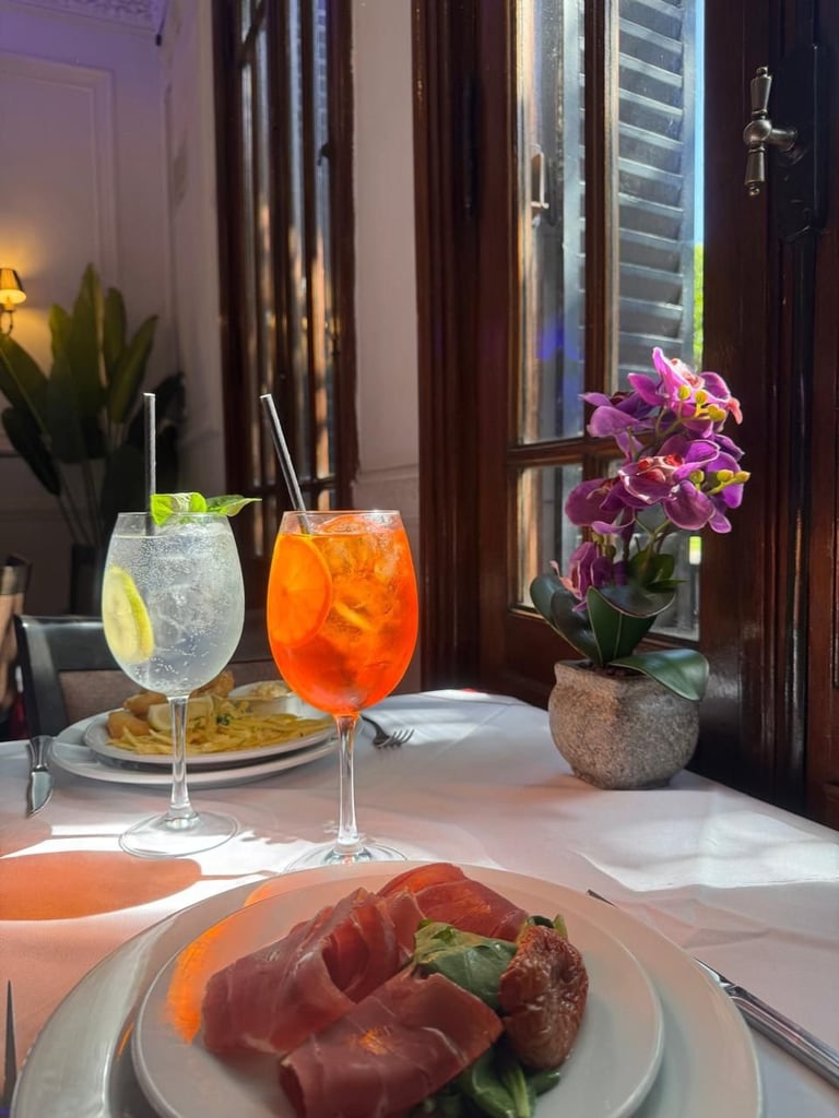 Fine dining table set with cocktails, cured meat appetizer, flowers, and city view through tall windows